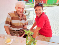 Ein Bewohner des Haus Elisabeth mit einer Betreuerin sitzen an einen Holztisch beim Weinfest.