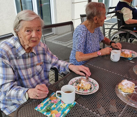 Zwei ältere Damen sitzen bei Tisch bei Kaffee und Kuchen. Dame links sieht ins Bild.