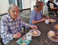 Zwei ältere Damen sitzen bei Tisch bei Kaffee und Kuchen. Dame links sieht ins Bild.