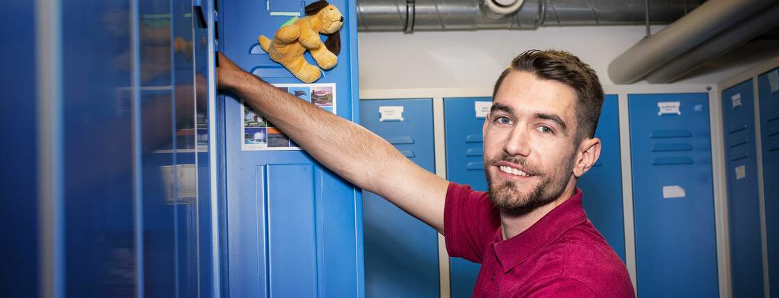 Ein junger Mann mit Bart und roten T-shirt mit Caritas Aufdruck holt Sachen aus einem blauen Spint