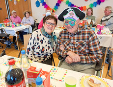 Ein Paar feiert fröhlich bei der Faschingsfeier im Haus Lisa. Der Mann trägt einen Piratenhut und eine bunte Maske, die Frau trägt weiße Katzenohren. Im Hintergrund sitzen weitere Gäste, umgeben von bunten Dekorationen.