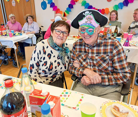 Ein Paar feiert fröhlich bei der Faschingsfeier im Haus Lisa. Der Mann trägt einen Piratenhut und eine bunte Maske, die Frau trägt weiße Katzenohren. Im Hintergrund sitzen weitere Gäste, umgeben von bunten Dekorationen.