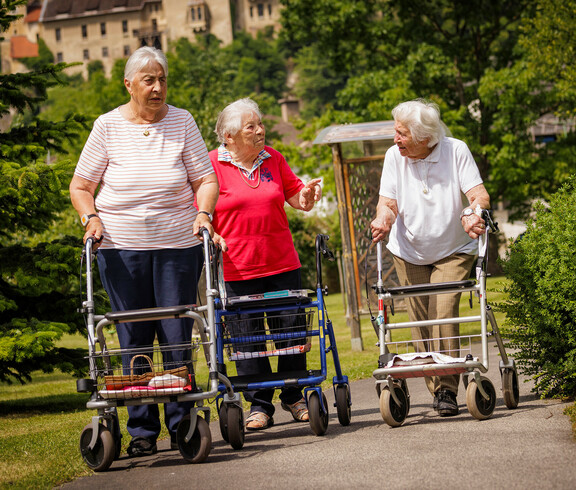 Drei Damen spazieren mit ihren Rollatoren durch den schönen Garten des Hauses.