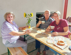 Besucher*innen des Erntedankfest im Haus Elisabeth sitzen gemeinsam an einem Holztisch im Freien und trinken Kaffee. Sie wirken entspannt und genießen die Gesellschaft. Auf dem Tisch stehen Sonnenblumen in einer Vase und einige Brezeln. Alle drei Personen lächeln freundlich in die Kamera.