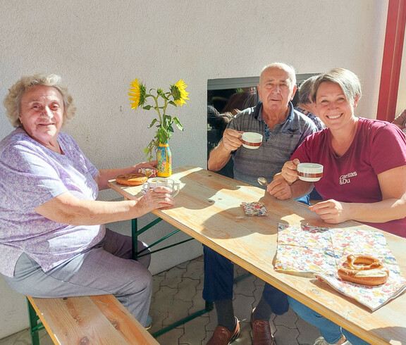 Besucher*innen des Erntedankfest im Haus Elisabeth sitzen gemeinsam an einem Holztisch im Freien und trinken Kaffee. Sie wirken entspannt und genießen die Gesellschaft. Auf dem Tisch stehen Sonnenblumen in einer Vase und einige Brezeln. Alle drei Personen lächeln freundlich in die Kamera.