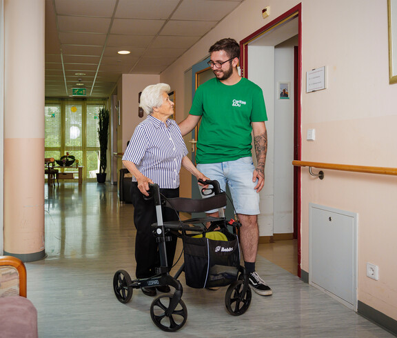 Eine Dame mit Rollator lächelt einem Pfleger zu, während sie im Gang des Pflegewohnhauses stehen.