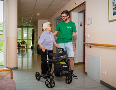 Eine Dame mit Rollator lächelt einem Pfleger zu, während sie im Gang des Pflegewohnhauses stehen.