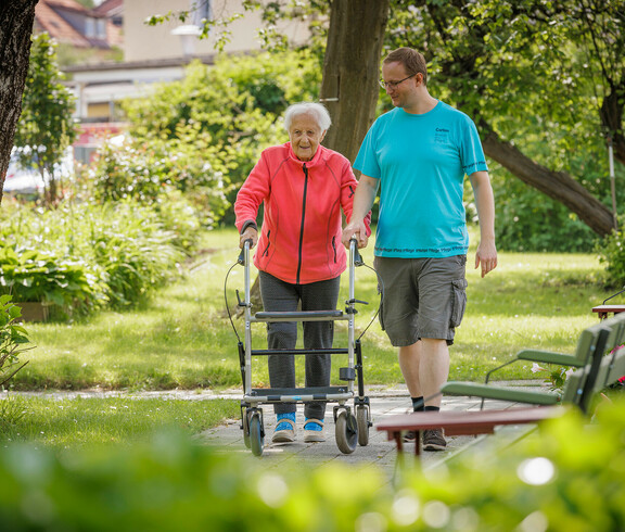 Eine ältere Dame begleitet von einem Pfleger gehen im Garten spazieren.