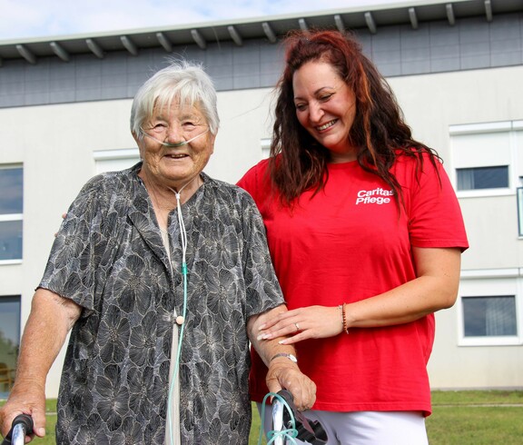 Eine Bewohnerin vom Haus Lisa, welche mit einem Gehwagen steht und Sauerstoffschläuche trägt. Neben ihr steht eine lächelnde Pflegekraft in einem roten T-Shirt mit der Aufschrift "Caritas Pflege", die sie unterstützend am Arm hält. Beide wirken zufrieden und fröhlich. 