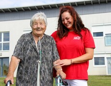 Eine Bewohnerin vom Haus Lisa, welche mit einem Gehwagen steht und Sauerstoffschläuche trägt. Neben ihr steht eine lächelnde Pflegekraft in einem roten T-Shirt mit der Aufschrift "Caritas Pflege", die sie unterstützend am Arm hält. Beide wirken zufrieden und fröhlich. 