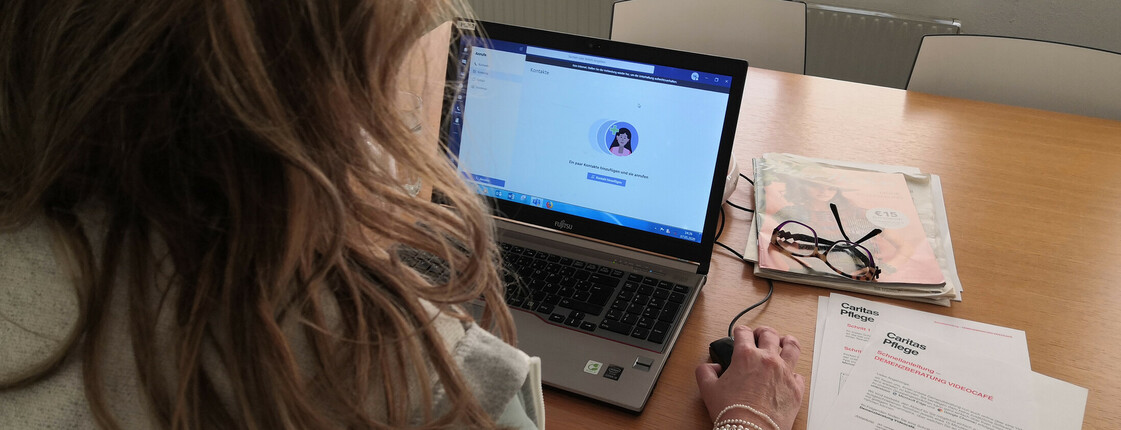 Eine Frau sitzt mit dem Rücken zur Kamera und sieht auf einem Laptopbildschirm, neben ihr liegt die Anleitung zum Einstieg in das "Demenzfrühstück" ein Angebot der Caritas für Pflegende Angehörige von Menschen mit Demenz.