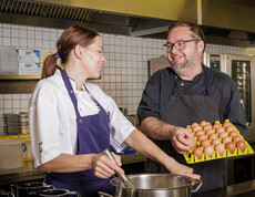 Eine Köchin ruht das Essen im Top, dabei sieht sie den Küchenchef an, der eine Lage Eier in der Hand hält. Beide lächeln sich an. 