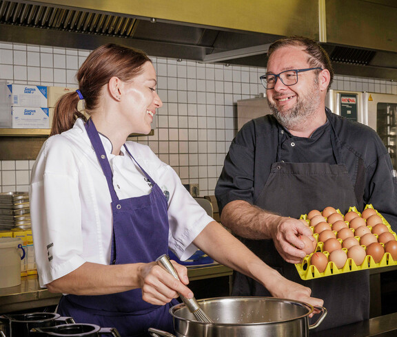 Eine Köchin ruht das Essen im Top, dabei sieht sie den Küchenchef an, der eine Lage Eier in der Hand hält. Beide lächeln sich an. 