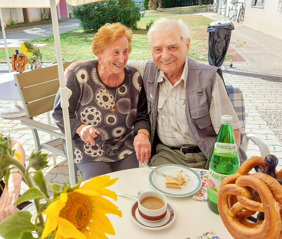 Zwei Personen genießen das Erntedankfest im Haus Elisabeth, indem sie gemeinsam an einen Tisch im Freien sitzen und in die Kamera lächeln. Vor ihnen stehen eine Tasse Kaffee, ein Stück Kuchen und eine Flasche Wasser. 