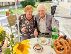 Zwei Personen genießen das Erntedankfest im Haus Elisabeth, indem sie gemeinsam an einen Tisch im Freien sitzen und in die Kamera lächeln. Vor ihnen stehen eine Tasse Kaffee, ein Stück Kuchen und eine Flasche Wasser. 