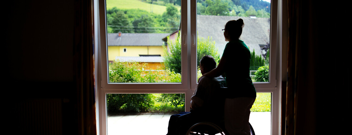 Eine Frau steht mit einem Menschen im Rollstuhl, die nur als Schatten gesehen werden, weil sie vor einem beleuchtenden Fenster stehen.