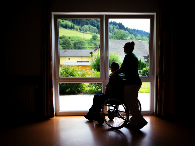 Eine Frau steht mit einem Menschen im Rollstuhl, die nur als Schatten gesehen werden, weil sie vor einem beleuchtenden Fenster stehen.