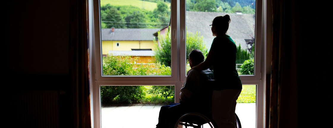 Eine Frau steht mit einem Menschen im Rollstuhl, die nur als Schatten gesehen werden, weil sie vor einem beleuchtenden Fenster stehen.