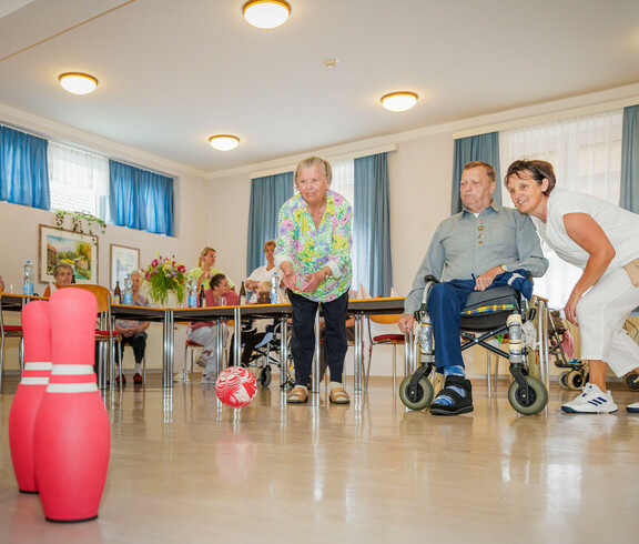 Eine Gruppe von Bewohner*innen sitzt im Gemeinschaftsraum um aufgestellte Kegel und versucht diese mit einem Ball umzustoßen. 