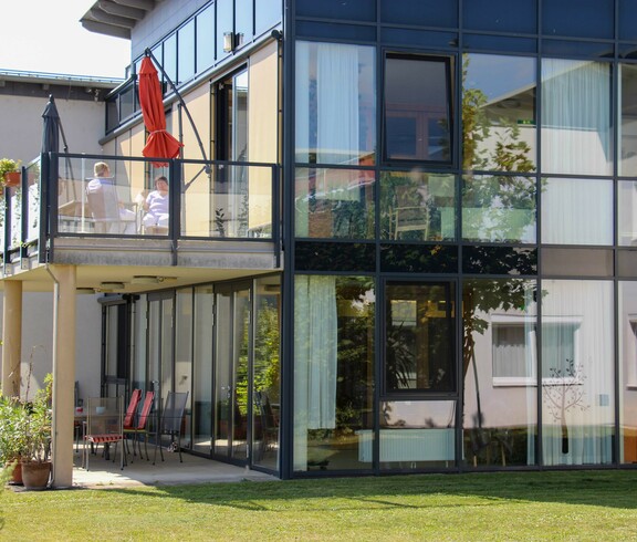 Außenansicht des Hauses Lisa mit großen Glasfronten und einem Balkon, auf dem zwei Personen sitzen. Zu sehen ist auch eine Grünfläche.