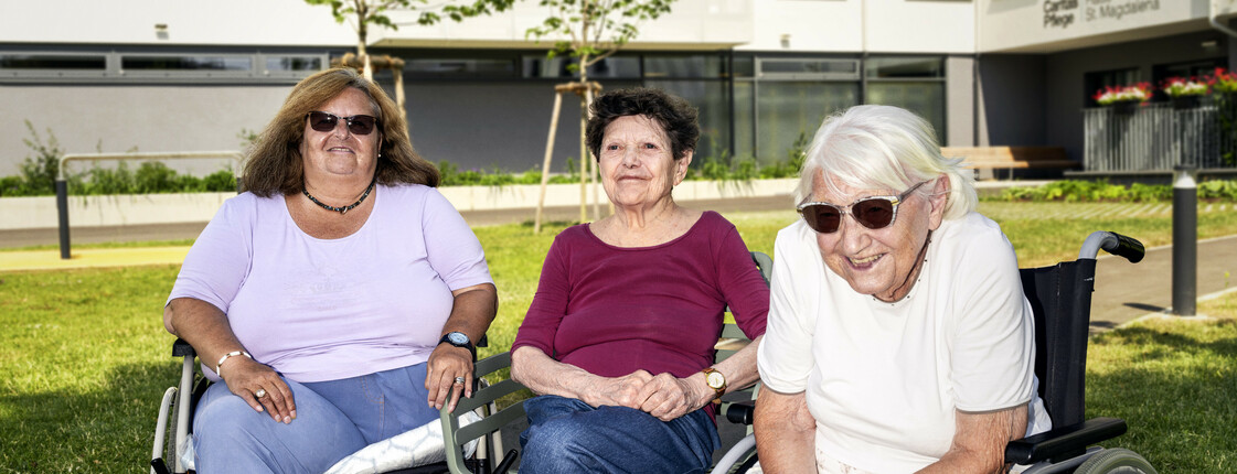 Drei ältere Frauen sitzen auf ihren Rollatoren bzw. Rollstühlen nebeneinander, sie lächeln in die Kamera. Im Hintergrund ist ein Pflegewohnhaus zu erkennen.