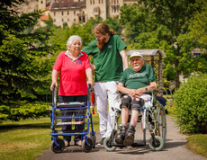 Ein Pfleger steht mit einer Dame am Rollator und einem Mann im Rollstuhl im Garten des Hauses und lächelt die Dame an.