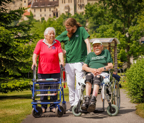 Ein Pfleger steht mit einer Dame am Rollator und einem Mann im Rollstuhl im Garten des Hauses und lächelt die Dame an.