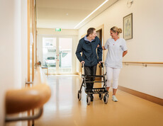 Eine Mitarbeiterin spaziert mit einem Bewohner am Rollator durch einen Gang unseres Pflegewohnhauses.