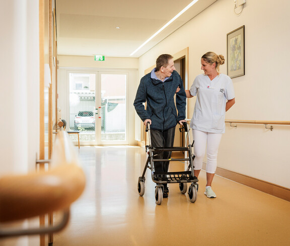 Eine Mitarbeiterin spaziert mit einem Bewohner am Rollator durch einen Gang unseres Pflegewohnhauses.