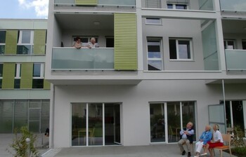 Ansicht des Hauses, auf einem Balkon im ersten Stock stehen zwei Frauen uns sehen auf zwei Frauen uns einen Mann herunter, die vor dem Haus auf einer Bank sitzen. 