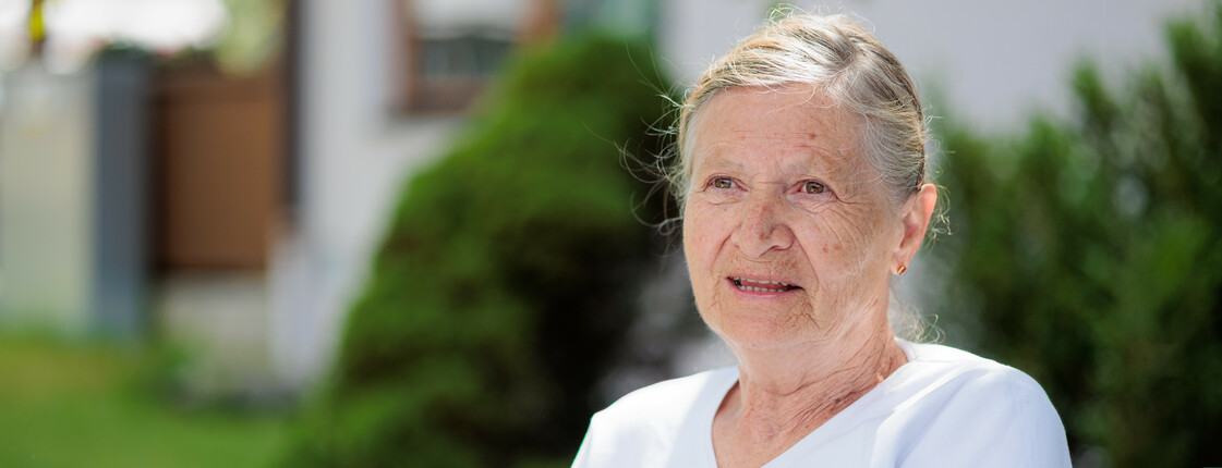 Eine Seniorin mit weißem T-Shirt sitzt im Sommer in einem grünen Garten.