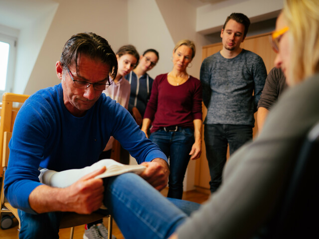 Ein Schüler der Schule für Sozialbetreuungsberufe zieht einer Mitschülerin einen Kompressionsstrumpf an um die zu üben. Seine restlichen Schüler*innen und die Lehrerin sehen dabei im Hintergrund zu.