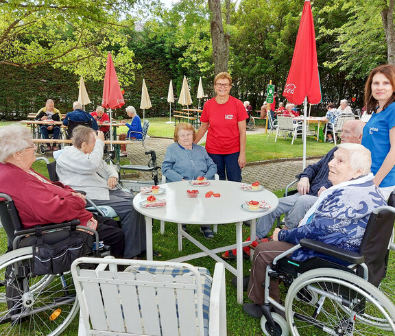 Man sieht die Bewohnerinnen des Haus Elisabeth mit Betreuerinnen. Sie genießen an einen Tisch im Freien das Erbeerfest. Es steht Erdbeerkuchen am Tisch