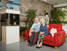 Ein Bewohner und eine Bewohnerin sitzen auf der gemütlichen roten Couch in unserem Aufenthaltsraum im Eingangsbereich, in dem auch die alte Stadtmauer von Klagenfurt integriert ist.