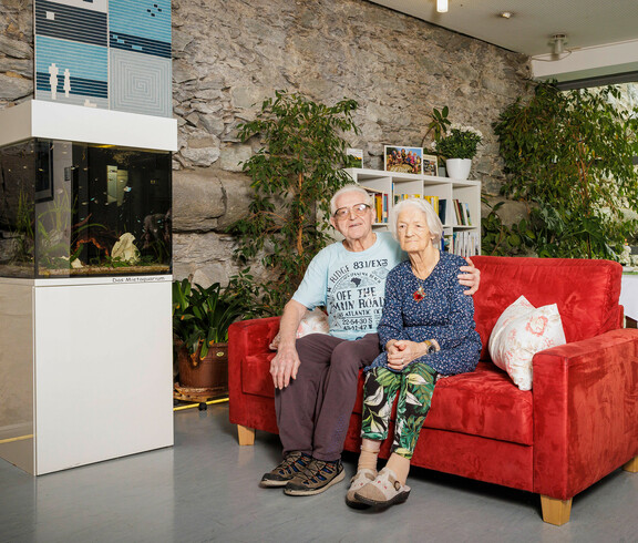 Ein Bewohner und eine Bewohnerin sitzen auf der gemütlichen roten Couch in unserem Aufenthaltsraum im Eingangsbereich, in dem auch die alte Stadtmauer von Klagenfurt integriert ist.