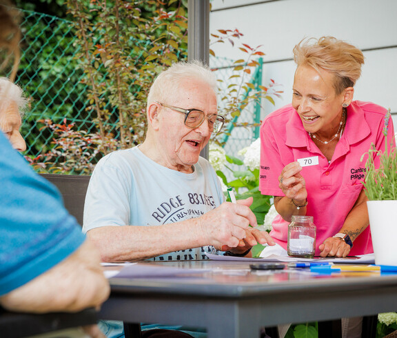 Eine Betreuerin hält einen Zettel mit der Zahl 70 hoch, ein Bewohner kreuzt auf seinem Bingo-Zettel diese Zahl ab.