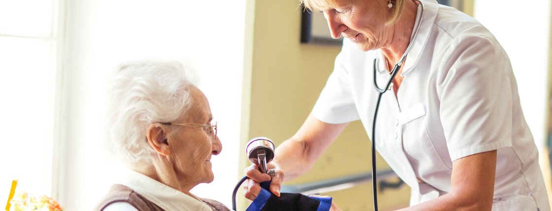 Eine Diplomierte Gesundheits- und Kranenpflegerin misst bei einer älteren Dame im Rollstuhl den Blutdruck.