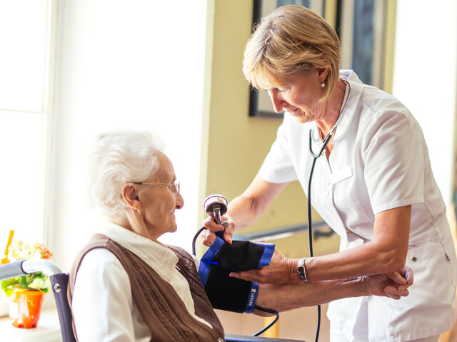 Eine Diplomierte Gesundheits- und Kranenpflegerin misst bei einer älteren Dame im Rollstuhl den Blutdruck.