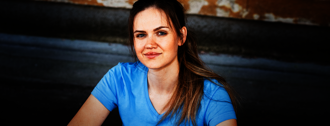 Eine junge Frau mit braunen langen Haaren und Pferdeschwanz sitzt auf einer Stiege, sie trägt ein hellblaues T-Shirt mit Caritas Logo und lächelt in die Kamera.