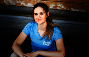 Eine junge Frau mit braunen langen Haaren und Pferdeschwanz sitzt auf einer Stiege, sie trägt ein hellblaues T-Shirt mit Caritas Logo und lächelt in die Kamera.