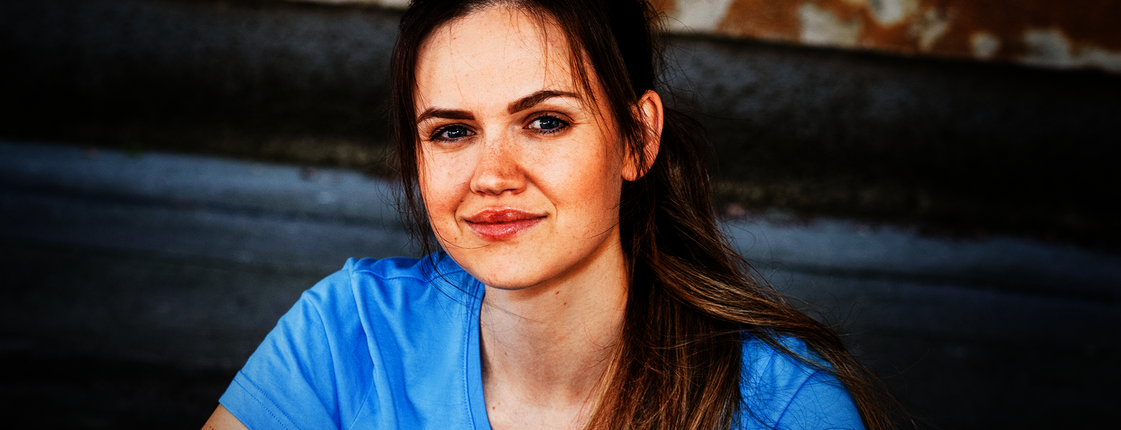 Eine junge Frau mit braunen langen Haaren und Pferdeschwanz sitzt auf einer Stiege, sie trägt ein hellblaues T-Shirt mit Caritas Logo und lächelt in die Kamera.