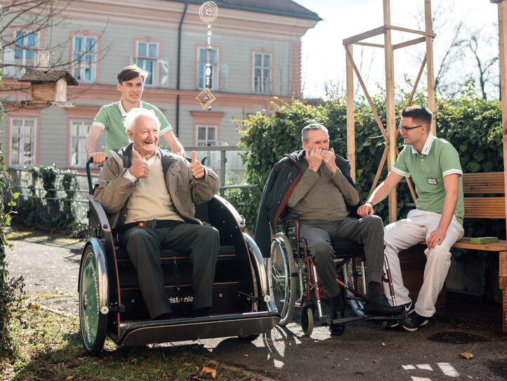 Ein Zivildiener fährt mit einem Bewohner in einer Rikscha durch den Garten, während ein zweiter Zivildiener mit einem Bewohner auf einer Bank sitzt und diesem beim Mundharmonika spielen zuhört.