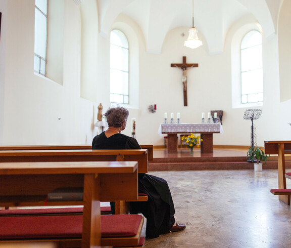 Eine ältere Dame sitzt in der hauseigenen Kapelle und blickt auf den Altar, auf dem sich 6 Kerzen und darunter ein Blumenstrauß befinden. Ein großes Christuskreuz hängt über dem Altar.