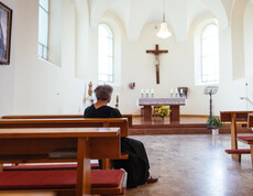 Eine ältere Dame sitzt in der hauseigenen Kapelle und blickt auf den Altar, auf dem sich 6 Kerzen und darunter ein Blumenstrauß befinden. Ein großes Christuskreuz hängt über dem Altar.