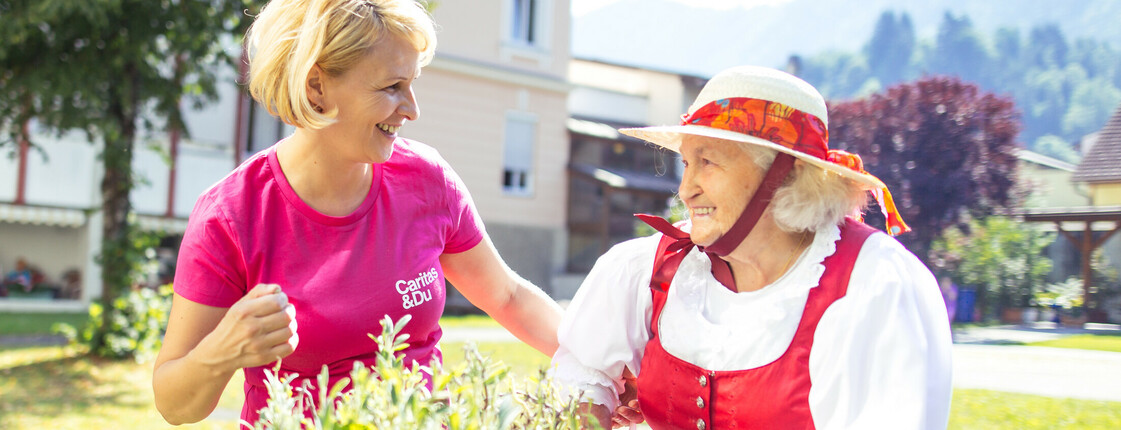 Eine junge Frau steht mit einer älteren Dame mit Gehstock an einem Hochbeet und die beiden lächeln sich an.