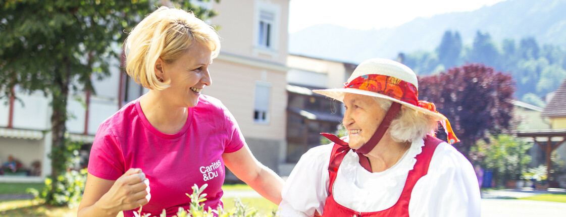 Eine junge Frau steht mit einer älteren Dame mit Gehstock an einem Hochbeet und die beiden lächeln sich an.