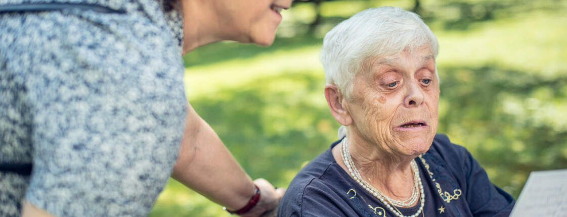 Eine Seniorin sitzt in einem Korbstuhl in einem Garten und liest eine Postkarte, welche sie in der Hand hochhält. Eine Pflegerin steht neben ihr und schaut ebenfalls die Postkarte an.