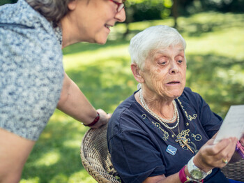 Eine Seniorin sitzt in einem Korbstuhl in einem Garten und liest eine Postkarte, welche sie in der Hand hochhält. Eine Pflegerin steht neben ihr und schaut ebenfalls die Postkarte an.