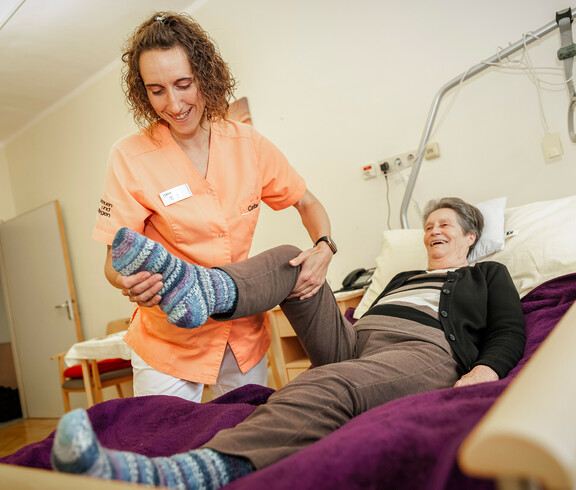 Eine Pflegekraft hilft einer Bewohnerin, die im Bett liegt mit Bewegungsübungen ihres Beines.