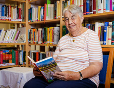 Eine ältere Dame sitzt vor einem Bücherregal, hat ein aufgeschlagenes Buch in der Hand und lächelt in die Kamera.